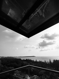 A black and white photo captures a coastal landscape. A portion of a building structure extends from the top left corner, creating a frame. Below, there is a glass balcony railing, and beyond it, a view of the sea with an island in the distance. The foreground includes a road with vehicles and lush green trees. The sky is partly cloudy, adding texture and depth to the scene.