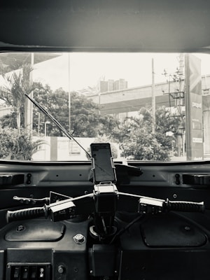 The image features the interior view from the driver's seat of an auto-rickshaw. The focus is on the handlebars and dashboard with a mounted mobile phone displaying a map. Through the windshield, a view of a city street with trees, a building, and a billboard is visible.