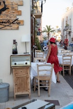 A street-side dining area features a wooden cabinet with an old-fashioned radio on top next to a table setting with wooden chairs and a white tablecloth. A person wearing a red dress and matching hat walks past the table. The street is lined with palm trees, and other tables are visible in the background, suggesting an outdoor dining atmosphere.