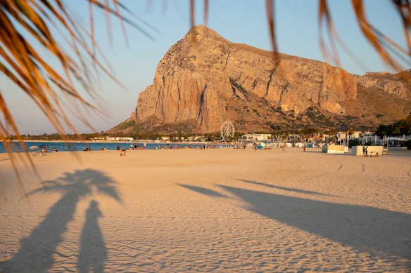 A sandy beach with scattered people enjoying leisure activities. A large, rugged mountain dominates the background, and lush greens are visible at its base. Palm tree shadows stretch across the sand, and a serene body of water lies adjacent to the shoreline.