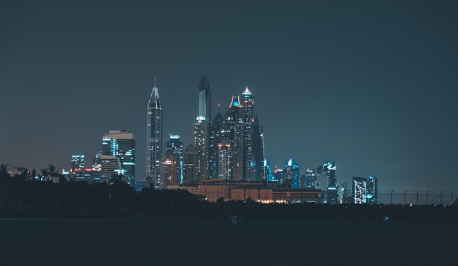 Skyline of a city at night with tall skyscrapers lit by various colored lights. The foreground is silhouetted with dark trees and a fence, adding to the urban nightscape.