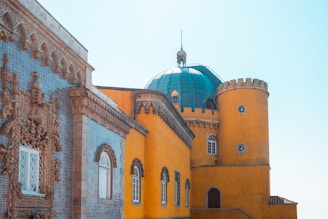 An architectural design featuring a striking blue-tiled facade with intricate stone carvings juxtaposed against a vibrant yellow wall. The building has a cylindrical yellow tower with small windows and a large teal dome with a spire on top. The design elements suggest a blend of classical and Moorish architectural styles.