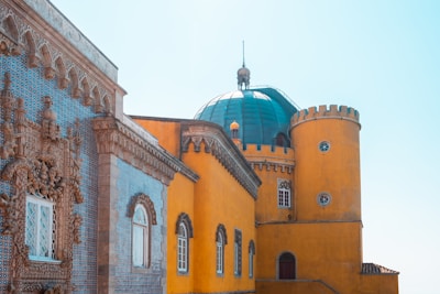 An architectural design featuring a striking blue-tiled facade with intricate stone carvings juxtaposed against a vibrant yellow wall. The building has a cylindrical yellow tower with small windows and a large teal dome with a spire on top. The design elements suggest a blend of classical and Moorish architectural styles.