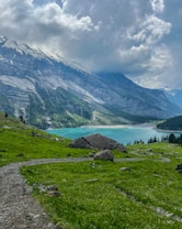 A serene landscape featuring a clear turquoise lake surrounded by dense forest at the base of a towering, rocky mountain range. The foreground includes a small grassy hill with wild green alpine meadows and a rock-strewn path leading to a rustic mountain hut.