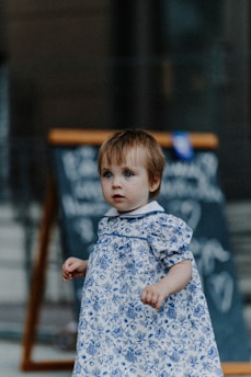 A young child with light brown hair is dressed in a white and blue floral patterned dress. The child stands in front of a blurred background that includes a chalkboard with unintelligible writing. The child has a curious and thoughtful expression.