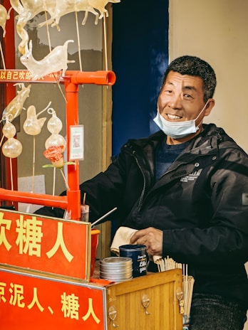 A man smiling while standing next to a stall that displays intricately crafted sugar candies in animal shapes. The stall is equipped with various tools and containers, with a prominent red sign featuring text in a foreign language.