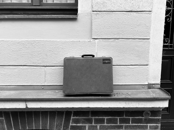 A black and white image featuring a solitary briefcase placed on an outdoor windowsill. The briefcase is centered against a textured stone wall divided by horizontal lines. Below the windowsill, there is brickwork and a small amount of graffiti on the metal surface.