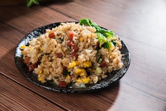 A black ceramic plate filled with fried rice featuring bits of vegetables, small pieces of cooked meat, and scrambled egg. The rice appears to be seasoned and garnished with green leafy vegetables, providing a colorful contrast against the wooden table surface.