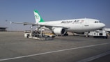 A large commercial airliner branded with the name Mahan Air is parked on an airport tarmac. The aircraft is being serviced with equipment such as a luggage cart nearby. The sky is clear, and the runway is expansive.
