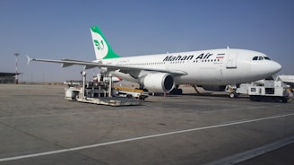 A large commercial airliner branded with the name Mahan Air is parked on an airport tarmac. The aircraft is being serviced with equipment such as a luggage cart nearby. The sky is clear, and the runway is expansive.
