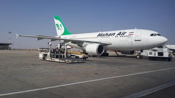 A large commercial airliner branded with the name Mahan Air is parked on an airport tarmac. The aircraft is being serviced with equipment such as a luggage cart nearby. The sky is clear, and the runway is expansive.