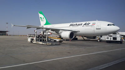 A large commercial airliner branded with the name Mahan Air is parked on an airport tarmac. The aircraft is being serviced with equipment such as a luggage cart nearby. The sky is clear, and the runway is expansive.