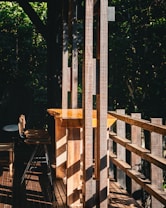 Sunlight filters through wooden slats creating shadows on a porch area. A wooden table with high chairs is positioned, suggesting a rustic outdoor setting. Lush greenery surrounds the space.