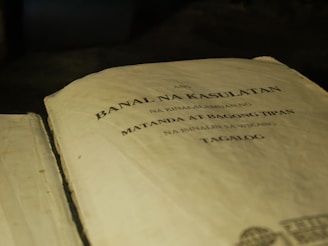 An open book with text in Tagalog, indicating it's a religious script, possibly a Bible. The page shows black printed letters on an aged, yellowish surface.