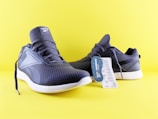A pair of dark blue athletic shoes with white soles is set against a bright yellow background. The shoes have a textured pattern and display a brand logo on the tongue. There is a tag attached to one of the shoes, providing product information.