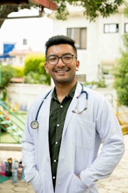 A person wearing a white lab coat and stethoscope stands outside in a sunny area. The individual is smiling and has short hair and glasses. The background features greenery, a building, and playground equipment.