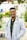A person wearing a white lab coat and stethoscope stands outside in a sunny area. The individual is smiling and has short hair and glasses. The background features greenery, a building, and playground equipment.