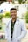 A person wearing a white lab coat and stethoscope stands outside in a sunny area. The individual is smiling and has short hair and glasses. The background features greenery, a building, and playground equipment.