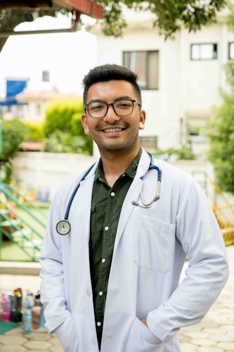 A person wearing a white lab coat and stethoscope stands outside in a sunny area. The individual is smiling and has short hair and glasses. The background features greenery, a building, and playground equipment.