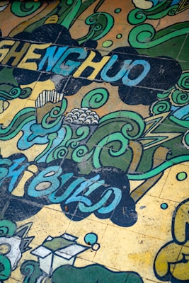 A colorful mural featuring swirling green and blue patterns interspersed with abstract shapes and words. The design includes cloud-like formations and stylized waves, with some small geometric elements. Letters forming words are incorporated into the flowing design.