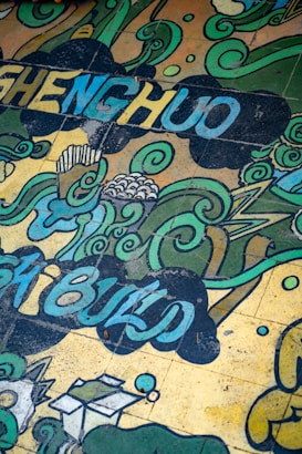 A colorful mural featuring swirling green and blue patterns interspersed with abstract shapes and words. The design includes cloud-like formations and stylized waves, with some small geometric elements. Letters forming words are incorporated into the flowing design.