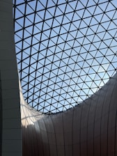 A geometrically intricate glass ceiling with a lattice structure, allowing natural light to illuminate the curving, metallic-clad walls below. The architectural design combines modern aesthetic with functional space.