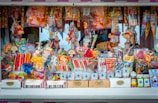 A colorful and vibrant candy stand filled with an assortment of sweets, including lollipops, candy sticks, gummies, and packaged treats. The display is densely packed with different shapes and sizes of candies in a variety of bright, eye-catching colors. Some toys and novelty items are also intermixed with the candies.