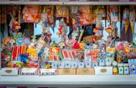 A colorful and vibrant candy stand filled with an assortment of sweets, including lollipops, candy sticks, gummies, and packaged treats. The display is densely packed with different shapes and sizes of candies in a variety of bright, eye-catching colors. Some toys and novelty items are also intermixed with the candies.