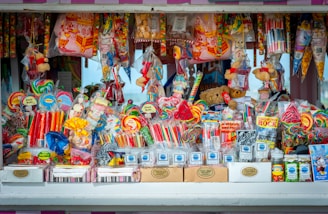A colorful and vibrant candy stand filled with an assortment of sweets, including lollipops, candy sticks, gummies, and packaged treats. The display is densely packed with different shapes and sizes of candies in a variety of bright, eye-catching colors. Some toys and novelty items are also intermixed with the candies.