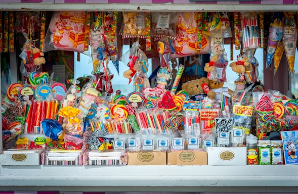 A colorful and vibrant candy stand filled with an assortment of sweets, including lollipops, candy sticks, gummies, and packaged treats. The display is densely packed with different shapes and sizes of candies in a variety of bright, eye-catching colors. Some toys and novelty items are also intermixed with the candies.