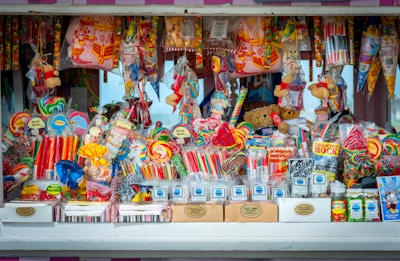 A colorful and vibrant candy stand filled with an assortment of sweets, including lollipops, candy sticks, gummies, and packaged treats. The display is densely packed with different shapes and sizes of candies in a variety of bright, eye-catching colors. Some toys and novelty items are also intermixed with the candies.