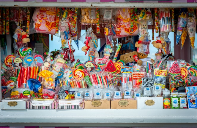 A colorful and vibrant candy stand filled with an assortment of sweets, including lollipops, candy sticks, gummies, and packaged treats. The display is densely packed with different shapes and sizes of candies in a variety of bright, eye-catching colors. Some toys and novelty items are also intermixed with the candies.
