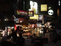 A vibrant night market scene with numerous food stalls illuminated by bright signs and lights. Several people are seen enjoying meals, talking, and preparing food. The atmosphere appears bustling and lively with a variety of colors from storefront signs.