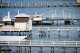 Wooden piers and small structures built over a calm body of water. The scene includes several docks extending into the blue water, with one featuring a small white cabin and another covered with a white canopy. The piers are supported by stilts and have white railings, and there are ladders and floating buoys visible in the water.