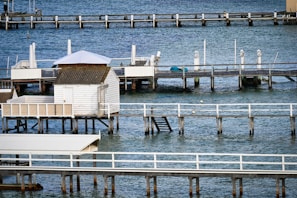 Wooden piers and small structures built over a calm body of water. The scene includes several docks extending into the blue water, with one featuring a small white cabin and another covered with a white canopy. The piers are supported by stilts and have white railings, and there are ladders and floating buoys visible in the water.