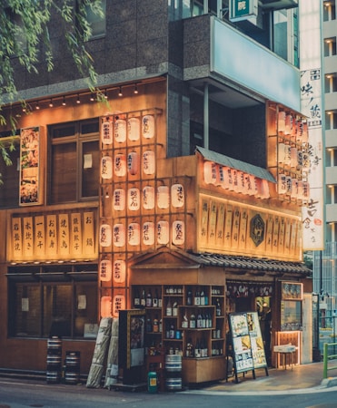 A vibrant street scene in an urban setting showcases a traditional Japanese izakaya, adorned with illuminated paper lanterns. The wooden facade is filled with Japanese characters, likely menu items or restaurant names. Shelves display a variety of sake bottles, and visual menus are placed outside to attract passersby. The warm glow of the lanterns contrasts with the surrounding modern architecture.