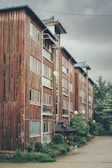 A multi-storey building with a rustic wooden exterior, featuring numerous windows and satellite dishes. The structure has a traditional Asian architectural element at the entrance, surrounded by lush greenery and a neatly maintained garden. The sky is overcast, giving a moody ambiance to the scene.