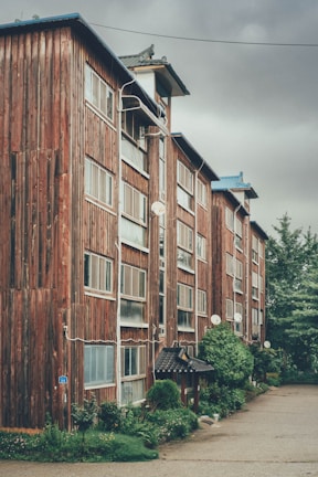 A multi-storey building with a rustic wooden exterior, featuring numerous windows and satellite dishes. The structure has a traditional Asian architectural element at the entrance, surrounded by lush greenery and a neatly maintained garden. The sky is overcast, giving a moody ambiance to the scene.