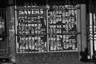 The image features a storefront window with painted signage advertising various products and prices, partially obscured by metal bars. The signage includes festive greetings and promotional offers for hampers and food items like eggs and milk. The numbers are large and bold, likely intended to catch the attention of passersby. There is also a partial view of a car at the bottom left corner.