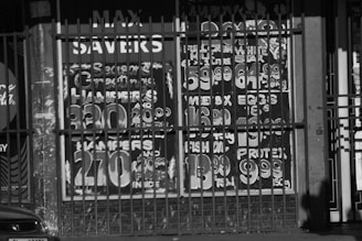The image features a storefront window with painted signage advertising various products and prices, partially obscured by metal bars. The signage includes festive greetings and promotional offers for hampers and food items like eggs and milk. The numbers are large and bold, likely intended to catch the attention of passersby. There is also a partial view of a car at the bottom left corner.