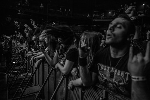 A black and white photo captures an energetic crowd at a concert, with people standing close to a barrier. Several individuals have their hands raised in rock gesture, while one person's hair is in motion, suggesting headbanging. The atmosphere is lively and intense, with many people wearing dark clothing and enthusiastic expressions.