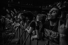 A black and white photo captures an energetic crowd at a concert, with people standing close to a barrier. Several individuals have their hands raised in rock gesture, while one person's hair is in motion, suggesting headbanging. The atmosphere is lively and intense, with many people wearing dark clothing and enthusiastic expressions.