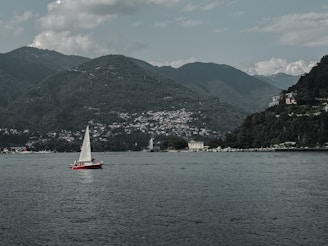A red sailboat with a white sail is on a calm body of water, with a picturesque backdrop of green mountains. There are several buildings nestled along the shoreline and into the hills, surrounded by dense vegetation. The sky is partly cloudy, adding to the serene and tranquil atmosphere.