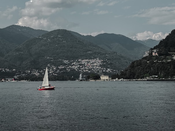 A red sailboat with a white sail is on a calm body of water, with a picturesque backdrop of green mountains. There are several buildings nestled along the shoreline and into the hills, surrounded by dense vegetation. The sky is partly cloudy, adding to the serene and tranquil atmosphere.