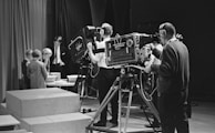 A black and white scene in a television studio features several people involved in a production. Multiple large vintage television cameras are operated by crew members wearing headsets. In the background, a group of children stand near a table looking at objects, possibly participating in a show. Curtains and studio equipment are visible, contributing to the classic broadcast environment.