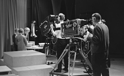 A black and white scene in a television studio features several people involved in a production. Multiple large vintage television cameras are operated by crew members wearing headsets. In the background, a group of children stand near a table looking at objects, possibly participating in a show. Curtains and studio equipment are visible, contributing to the classic broadcast environment.