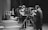 A black and white scene in a television studio features several people involved in a production. Multiple large vintage television cameras are operated by crew members wearing headsets. In the background, a group of children stand near a table looking at objects, possibly participating in a show. Curtains and studio equipment are visible, contributing to the classic broadcast environment.