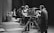 A black and white scene in a television studio features several people involved in a production. Multiple large vintage television cameras are operated by crew members wearing headsets. In the background, a group of children stand near a table looking at objects, possibly participating in a show. Curtains and studio equipment are visible, contributing to the classic broadcast environment.