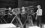 Three individuals operate large television cameras, each wearing headphones and focused on their tasks. The setting appears to be a television studio with multiple cameras on tripods. The individuals are standing behind the equipment, seemingly ready to capture a broadcast or production.