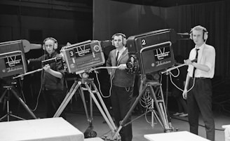 Three individuals operate large television cameras, each wearing headphones and focused on their tasks. The setting appears to be a television studio with multiple cameras on tripods. The individuals are standing behind the equipment, seemingly ready to capture a broadcast or production.